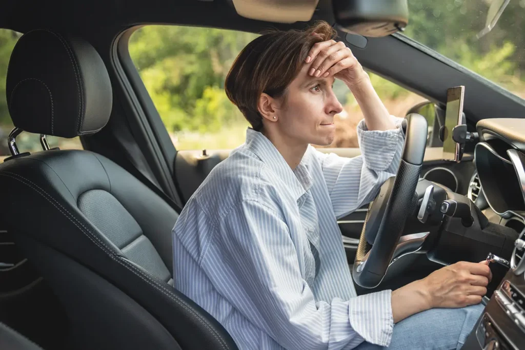 En person som sitter i førersetet i en bil med en hånd i pannen og ser ut av vinduet med et ettertenksomt uttrykk. Bilen står parkert, og personen, som ser ut til å reflektere over tillit i arbeidslivet, er iført stripet skjorte.