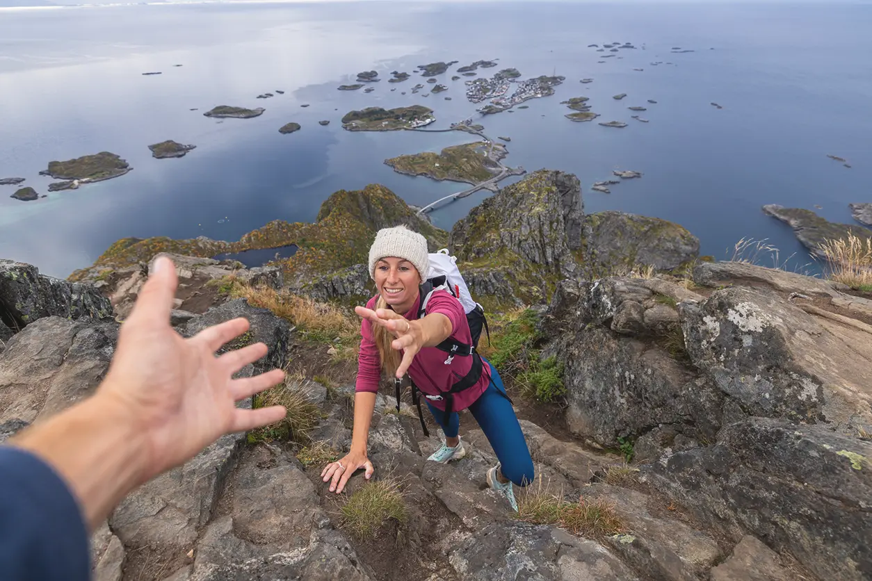 En person i turantrekk strekker seg ut til en annen persons hånd mens han klatrer et steinete fjell, med vidstrakt utsikt over kystøyene og vann i bakgrunnen.