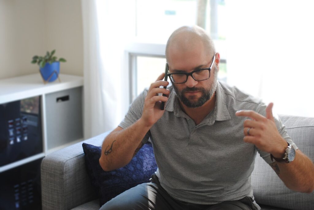 En mann med briller og skjegg sitter på en grå sofa, snakker i telefon om tillit i arbeidslivet, og gestikulerer med den andre hånden. En liten potteplante og et vindu er i bakgrunnen.