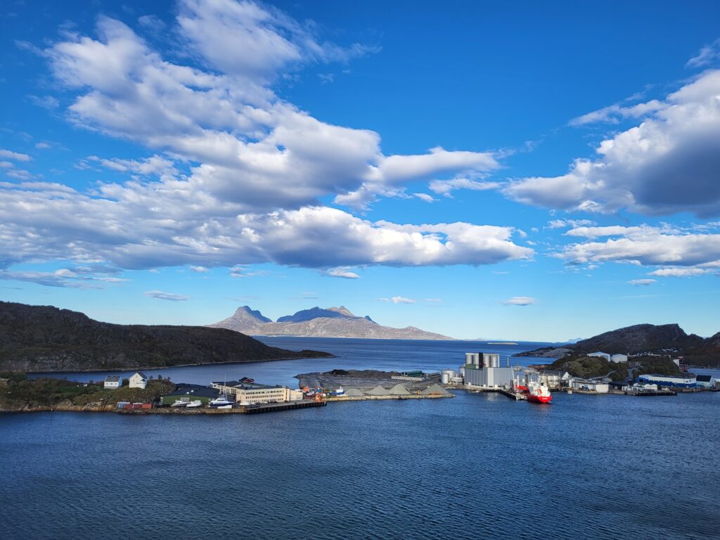 Et kystnært industriområde med bygninger og skip er satt i bakgrunn av åser og en delvis overskyet himmel over stille vann, noe som gjenspeiler tilliten til arbeidslivet som gjør at dette travle knutepunktet trives jevnt.