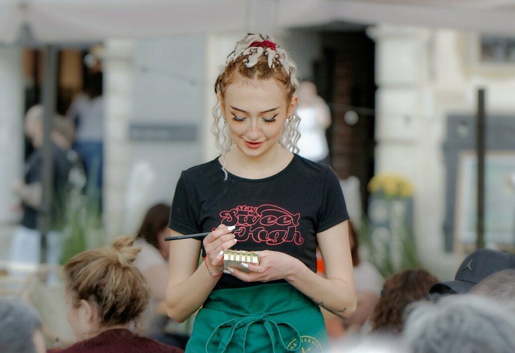 En kvinne iført en svart "Sweet Dough"-t-skjorte og grønt forkle skriver på en notisblokk, som legemliggjør *tillit i arbeidslivet*, mens hun står i en utendørs setting med folk i bakgrunnen.